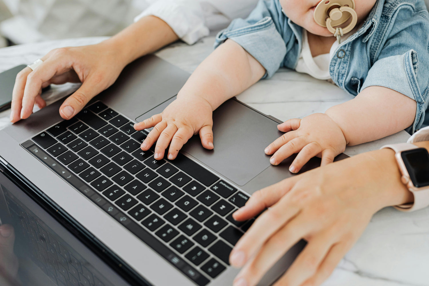 Baby with a pacifier playing with a laptop while an adult uses it